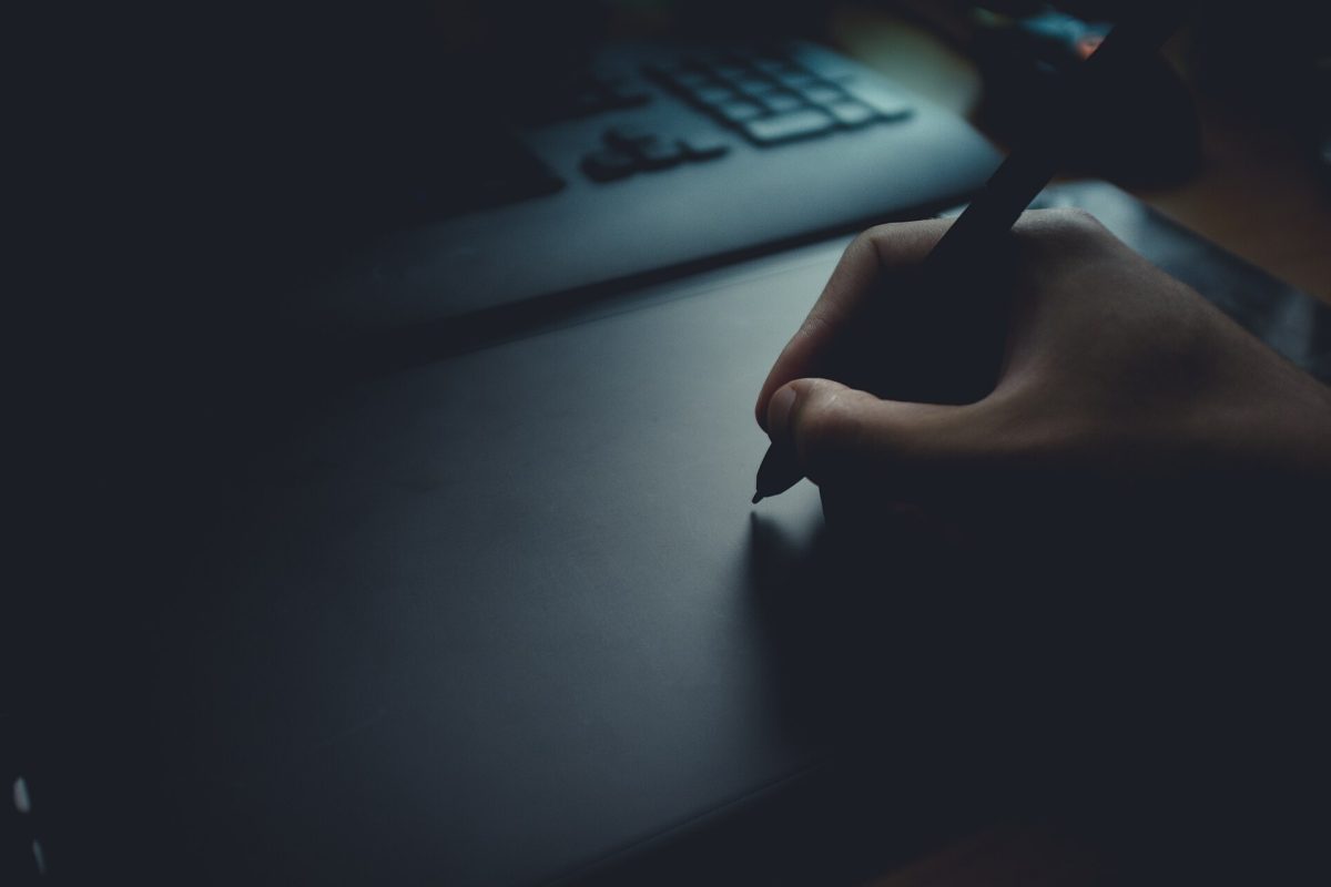 Hand holding a stylus on a graphics tablet, laptop keyboard blurred in the background