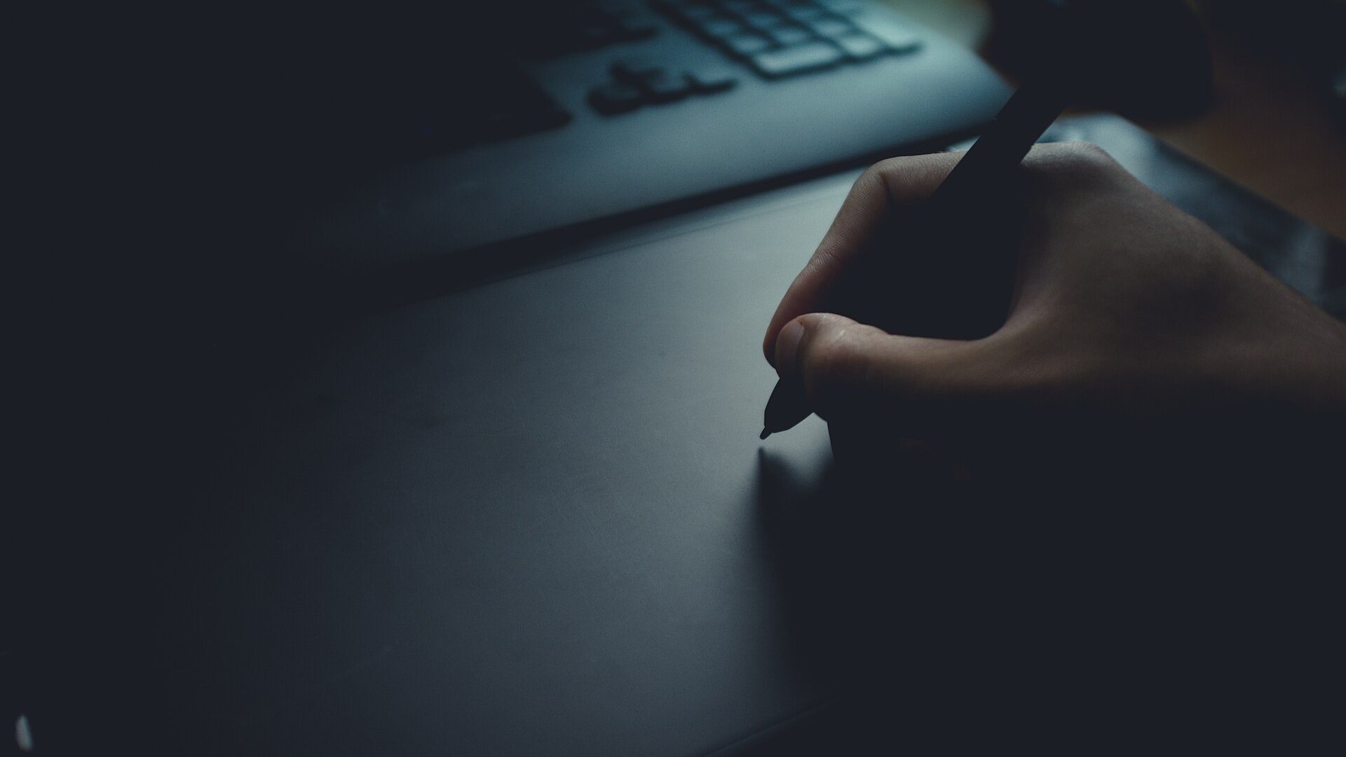 Hand holding a stylus on a graphics tablet, laptop keyboard blurred in the background