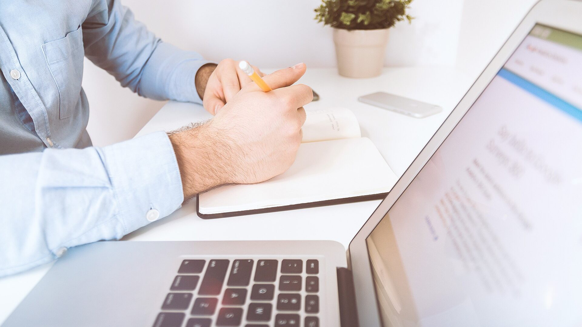 A person at a desk writing in a notebook beside an open laptop
